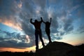 Silhouettes of man and woman on top of mountain. Hikers couple with raised arms on dramatic sky at sunset background.