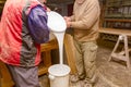 Two carpentry workers pour wood glue from a large plastic bucket into another empty