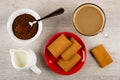 Spoon in bowl with instant coffee, cup of coffee with milk, jug of milk, cookies with filling in red saucer on table. Top view