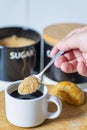 A teaspoon full of demerara sugar,being put into into a small,white cup of hot espresso coffee,in a domestic kitchen,