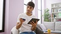 Tech-savvy young hispanic man relaxing on sofa, shopping online with credit card and touchpad indoors - portrait of serious but Royalty Free Stock Photo