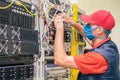 A technician in a medical mask switches wires in the server room. A man in a red cap works in a datacenter. A technical employee Royalty Free Stock Photo