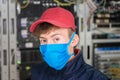 A technician in a red cap and medical mask is in the server room. The specialist is in the datacenter during the quarantine.
