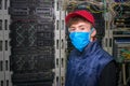 A technician in a red cap and medical mask is in the server room. The specialist is in the datacenter during the quarantine.