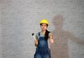 Technician woman ware yellow helmet with grey T-shirt and denim jeans apron dress standing with rubber hammer in hand and thumb up