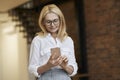 Technology. Beautiful mature business woman with blonde hair and glasses using her smartphone, standing indoors Royalty Free Stock Photo