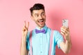 Technology concept. Funny man with moustache and bow-tie taking selfie on smartphone, showing peace sign and smiling at Royalty Free Stock Photo