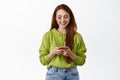 Technology. Smiling redhead girl looking at smartphone, chatting on phone, using application on cellphone, standing in