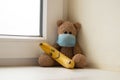 Teddy bear in a medical mask and next to a banana with a painted smile on the window in the apartment, childhood morbidity