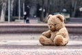 A teddy bear in the park sits on an idle fountain, a woman with a stroller leaves along the park alley.