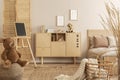 Teddy bear on pouf and small blackboard on easel in stylish kid`s bedroom with wooden cabinet and beige bedding on bed