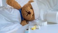 Teddy bear is sick in bed high fever and has a fever reducing sheet on the forehead. Beside the bed were thermometer and