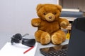 A teddy bear sits on the table, next to the laptop and headphones, a stack of sheets of paper in the background of shelves of book