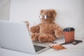 Teddy bear working concept. A cute teddy bear working in bed with a grey laptop and a cup of hot coffee