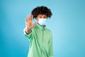 Teen black guy in face mask gesturing STOP, expressing negativity towards coronavirus pandemicon, blue background,