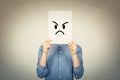 Teen boy covering face using a white paper sheet with angry sad emoticon sketch, like fake mask for hiding the real emotion from