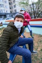 Teen boy and girl sitting on a seesaw on playground near high-rise buildings with apartments, a medical mask on their faces