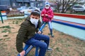 Teen boy and girl sitting on a seesaw on playground near high-rise buildings with apartments, a medical mask on their faces