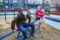 Teen boy and girl sitting on a seesaw on playground near high-rise buildings with apartments, a medical mask on their faces