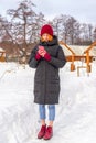 Teen cute girl in black down jacket, blue cropped jeans, burgundy hat, boots and fingerless gloves standing outdoor against winter Royalty Free Stock Photo