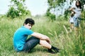 A cute teen with dark hair sitting on the grass embracing his legs. Mother is behind him. A teenager has difficult relationships