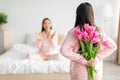 Teen girl hold bouquet of tulips behind back and congratulates asian millennial woman in bedroom interior