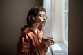 Teen girl nerd in glasses looking at window drinking cup of tea at home. Introvert, friendless