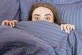Teen girl with open eyes covers her face with blue blanket in the morning. Beautiful woman lying down in the bed and sleeping Royalty Free Stock Photo