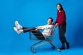 Teen girl pushing her teenage boyfriend in a shopping cart against blue background in studio Royalty Free Stock Photo