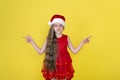Teen girl in red dress and Santa Claus red hat standing on yellow background shows direction. Girl in Santa hat isolated Royalty Free Stock Photo