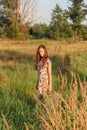 A young girl in a retro vintage dress with flowing hair is standing in a tall grass in a sunlight afterglow with a suitcase