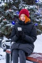 Teen girl sitting on bench in park against decorated fir tree in winter evening. Beautiful young woman wearing casual clothes Royalty Free Stock Photo