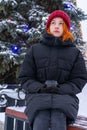 Teen girl sitting on bench in park against decorated fir tree in winter evening. Beautiful young woman wearing casual clothes Royalty Free Stock Photo