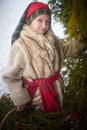 Teen girl in thick coat and a red sash with basket of fir branches and berries in cold winter day in forest. Medieval