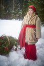 Teen girl in thick coat and a red sash with basket of fir branches and berries in cold winter day in forest. Medieval