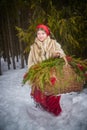 Teen girl in thick coat and a red sash with basket of fir branches and berries in cold winter day in forest. Medieval