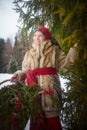 Teen girl in thick coat and a red sash with basket of fir branches and berries in cold winter day in forest. Medieval