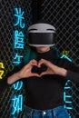 Teen girl using vr helmet shows heart gesture with hands on futuristic colorful background. Virtual love concept.