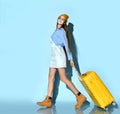 Teen lady in jeans overall, sweatshirt, sunglasses, boots and hat. Smiling, walking with yellow suitcase against blue background. Royalty Free Stock Photo