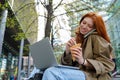 Teen redhead hipster girl using laptop talking on phone on city urban street. Royalty Free Stock Photo