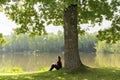 Teen young girl woman sitting on grass under huge high big old oak,trunk tree near lake,river, meditating,relaxing and smiling in Royalty Free Stock Photo