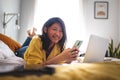 Teenage asian girl college student using mobile phone lying on bed while doing homework using laptop looking at camera. Royalty Free Stock Photo
