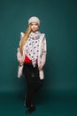 A teenage blonde model girl in modish winter clothes posing over the dark background in full length. Happy girl wearing