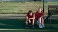 Teenage boy and girl in sports uniforms sit on football pitch near bench at sunlight