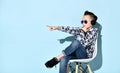 Teenage boy in headphones, sunglasses and casual clothes. Pointing at something, sitting on white chair against blue background Royalty Free Stock Photo