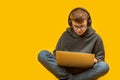 Teenage boy in jeans hoodie glasses and headphones sits cross-legged holding laptop. Online learning. Programming for children.