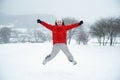 Teenage boy in red puffy jacket walks during snowfall. Child jumps up with his hands up and enjoys the winter holidays