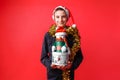 Teenage boy in Santa hat and tinsel on neck holding Christmas gift box on red background Royalty Free Stock Photo