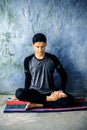 Teenage boy wearing black colored attire and doing yoga on colorful traditional mat and doing Fire Log Pose Agni Stambhasana wit