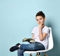 Teenage boy in white t-shirt, jeans and sneakers. He looking thoughtful, holding cash, sitting on white chair over blue background